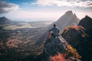 A hiker sits on a mountaintop, taking in a breathtaking view of a scenic valley and distant peaks.